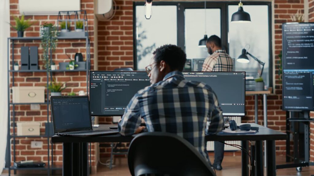 Programmer writes code on a laptop while sitting at a table, rear view