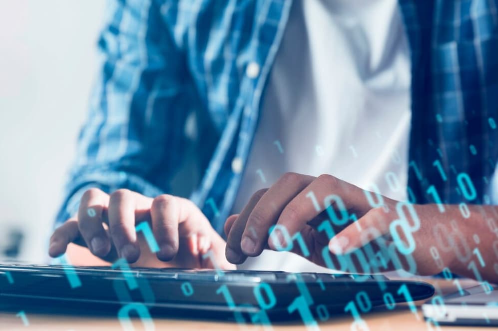 Close-up of hands typing on a keyboard with digital binary code overlay