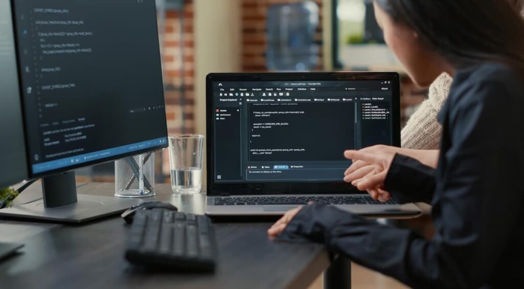 A woman is coding on a laptop with a second monitor on a wooden desk
