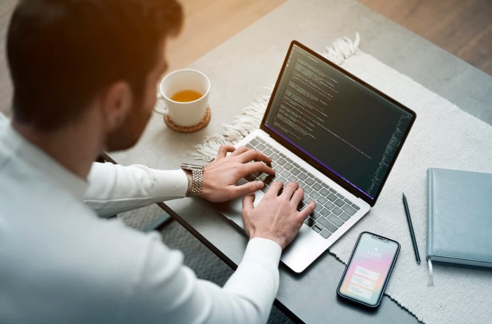 A man programs on a laptop with a phone and tea beside him